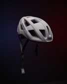 A white bicycle helmet is suspended in mid-air against a dark, gradient background, which transitions from blue to red. The helmet features several large ventilation holes and an adjustable chin strap. The brand name 'SMITH' is visible on the side.