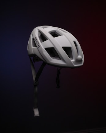 A white bicycle helmet is suspended in mid-air against a dark, gradient background, which transitions from blue to red. The helmet features several large ventilation holes and an adjustable chin strap. The brand name 'SMITH' is visible on the side.