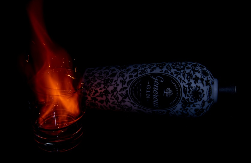 A glass with a vibrant orange flame emanating from it is positioned near a decorative gin bottle adorned with intricate patterns on a dark background. The bottle lies horizontally, partially obscured by shadow, highlighting the contrast between the bright flame and the bottle's dark patterns.