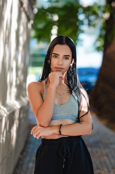 A woman with long dark hair is standing outdoors, wearing a gray sports bra and black pants. She has her left hand resting under her chin, and she gazes directly at the camera. The background includes tree foliage and a blurred pathway, creating a serene and peaceful setting.