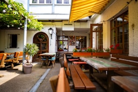 An outdoor seating area with wooden benches and tables arranged under a yellow-striped awning. Potted plants and red flowers in window boxes add a touch of greenery and color. The setting appears to be cozy, with sunlight filtering through, creating a warm and inviting atmosphere.