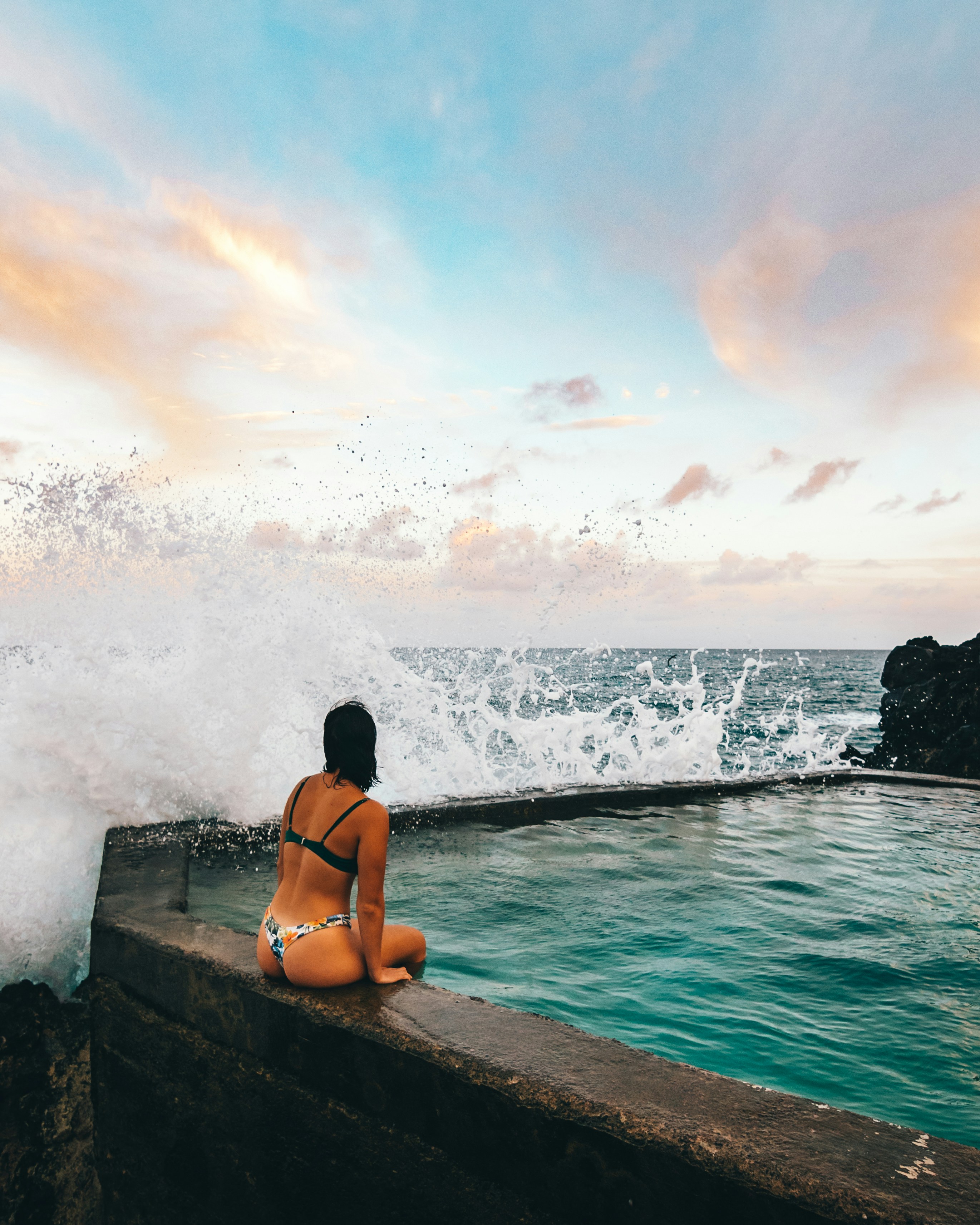 woman in black bikini sitting on rock near sea waves during daytime
