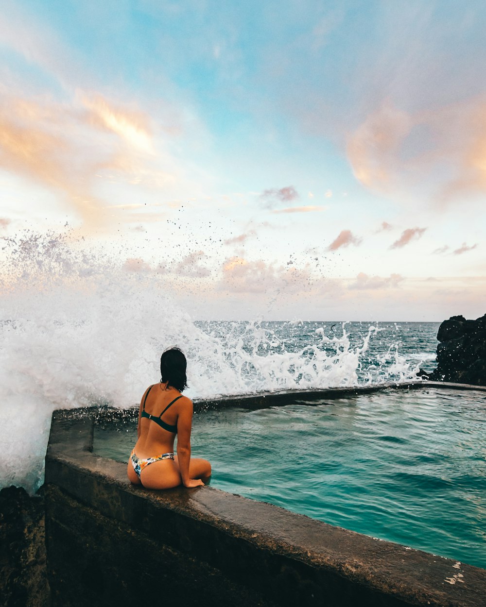 woman in black bikini sitting on rock near sea waves during daytime