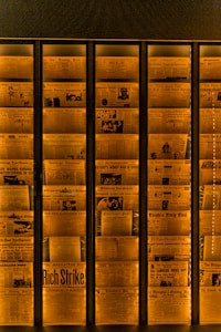 A display of vintage newspaper front pages arranged in a vertical alignment, illuminated with a warm orange light. Headlines with bold and varied fonts are visible, featuring historical news stories and photographs, giving an archival and historical feel.