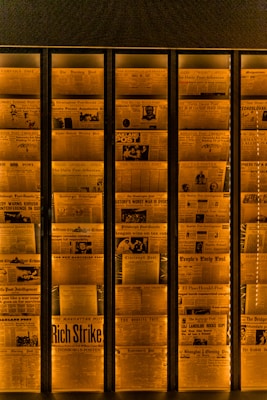 A display of vintage newspaper front pages arranged in a vertical alignment, illuminated with a warm orange light. Headlines with bold and varied fonts are visible, featuring historical news stories and photographs, giving an archival and historical feel.