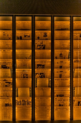 A display of vintage newspaper front pages arranged in a vertical alignment, illuminated with a warm orange light. Headlines with bold and varied fonts are visible, featuring historical news stories and photographs, giving an archival and historical feel.
