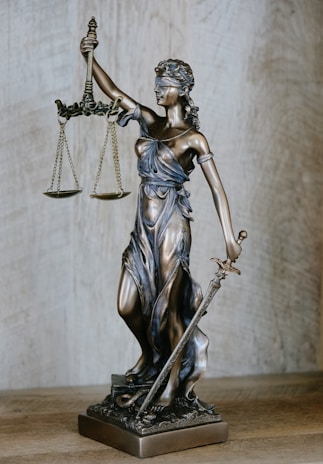 A bronze sculpture of Lady Justice, depicted with a blindfold, holding scales in one hand and a sword in the other. The figure stands on a detailed base featuring a snake and other ornate elements.