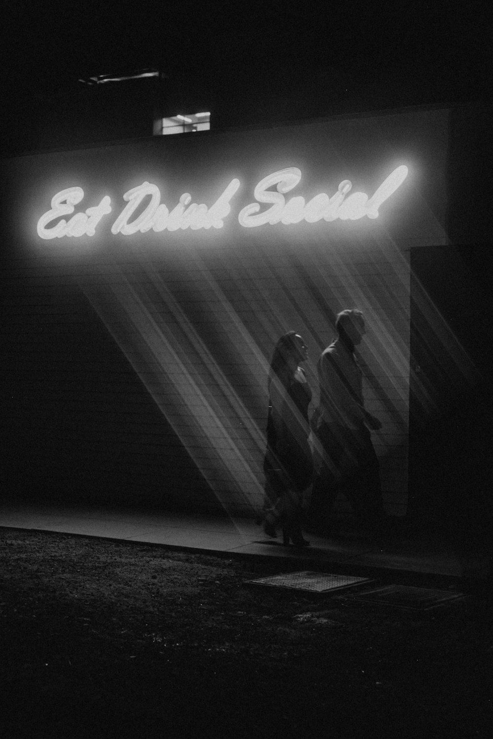 grayscale photo of man and woman standing in front of the UNKs neon light signage