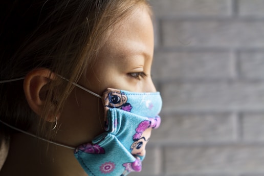 A child is facing sideways, wearing a colorful face mask that features cartoon designs. The background consists of a blurred, gray brick wall.