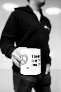 A person wearing a dark hoodie holds a white mug with partial text and a pretzel design printed on it. The image is in black and white, with a blurred background, focusing on the hand and the mug.