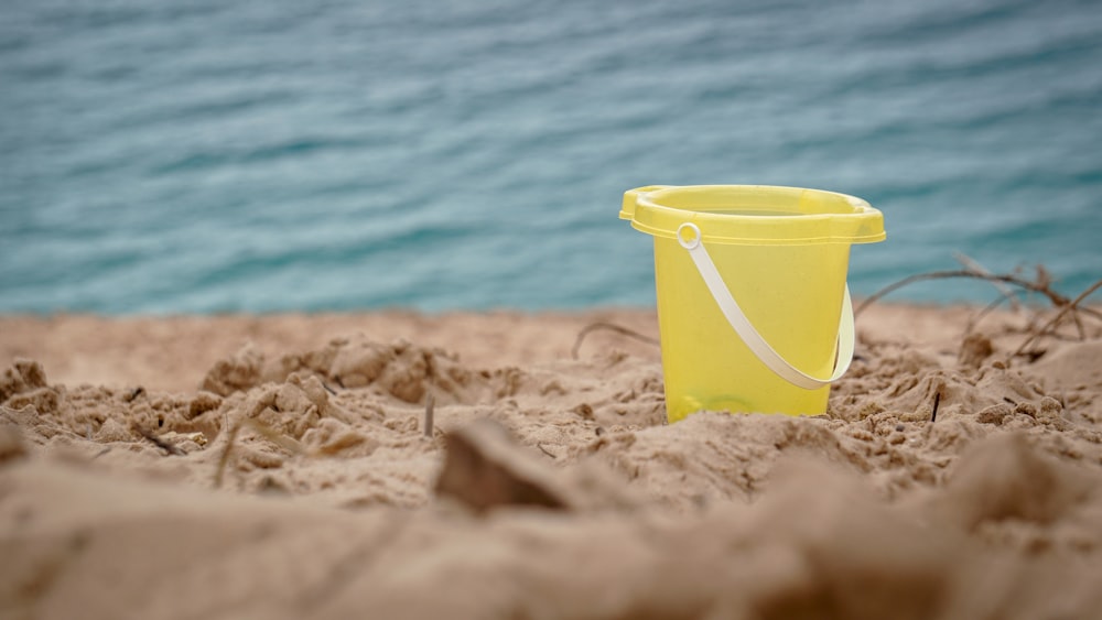 balde de plástico amarelo na areia marrom perto do corpo de água durante o dia