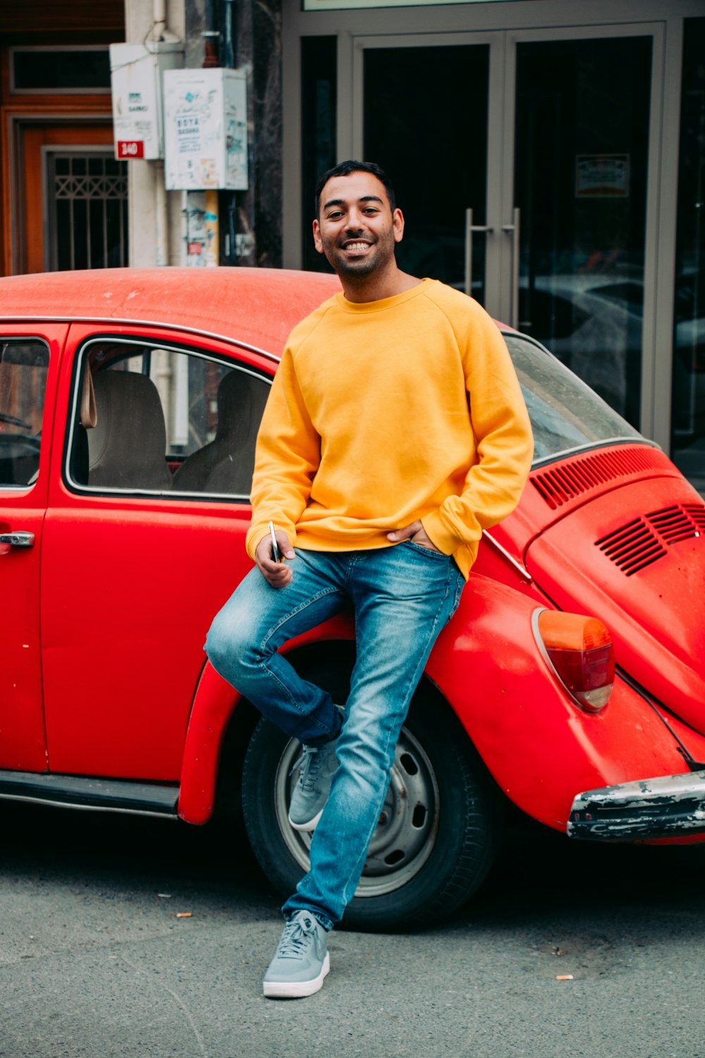 Mann in gelbem Pullover und blauer Jeans sitzt auf rotem Volkswagen Käfer