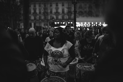 A group of people gathered outdoors, engaging in a drumming performance. The central figure is a woman holding drumsticks, surrounded by others with drums. The scene is captured in black and white, giving it a timeless and focused atmosphere.