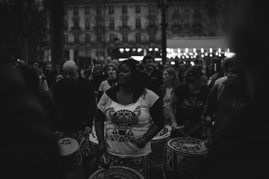 A group of people gathered outdoors, engaging in a drumming performance. The central figure is a woman holding drumsticks, surrounded by others with drums. The scene is captured in black and white, giving it a timeless and focused atmosphere.