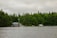 A serene lakeside scene featuring a rustic wooden cabin with a blue roof and a flagpole near the water's edge. The cabin is surrounded by dense, lush green forest, and the water is calm under an overcast sky. A dock extends into the lake with a small boat and a larger white boat moored nearby.