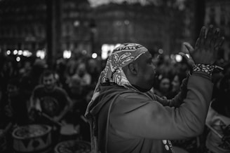 A scene with a group of people engaged in music and drumming, featuring a man in the foreground wearing a patterned headscarf and a sweatshirt, clapping his hands. The background is filled with a crowd in a dimly lit environment, indicating a lively atmosphere.