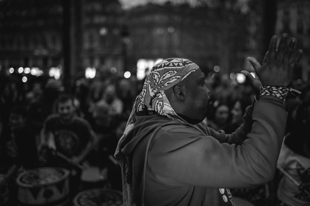 A scene with a group of people engaged in music and drumming, featuring a man in the foreground wearing a patterned headscarf and a sweatshirt, clapping his hands. The background is filled with a crowd in a dimly lit environment, indicating a lively atmosphere.