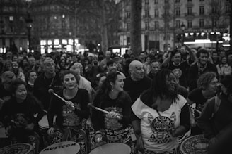A lively group of people participate in an outdoor drumming event. The scene is bustling with energy as the drummers, wearing patterned shirts, play their instruments enthusiastically. The crowd in the background adds to the energetic atmosphere, and the setting is urban, with tall buildings visible.