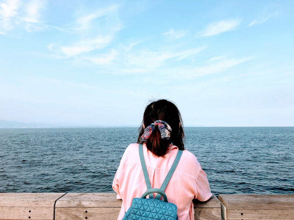 Donna in camicia rosa che si siede sulla panchina di legno marrone che guarda il mare durante il giorno