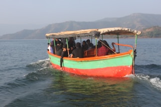 A small motorboat with a group of people on board is navigating through a calm body of water. The boat is painted with bright colors, including green, red, and orange. The passengers are seated under a canopy and appear to be enjoying a leisurely ride. In the background, there are distant hills under a clear sky.