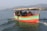 A small motorboat with a group of people on board is navigating through a calm body of water. The boat is painted with bright colors, including green, red, and orange. The passengers are seated under a canopy and appear to be enjoying a leisurely ride. In the background, there are distant hills under a clear sky.