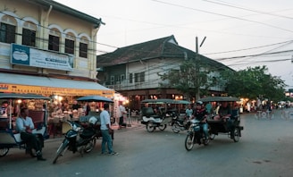 A busy street scene in an Asian city with several tuk-tuks and motorcycles parked and moving. On the left, there is an art shop displaying various colorful items under a yellow sign. Many people are gathered around, with some appearing as tourists and others as locals. Buildings with traditional architecture and a mix of old and new styles surround the street.