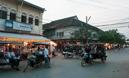 A busy street scene in an Asian city with several tuk-tuks and motorcycles parked and moving. On the left, there is an art shop displaying various colorful items under a yellow sign. Many people are gathered around, with some appearing as tourists and others as locals. Buildings with traditional architecture and a mix of old and new styles surround the street.