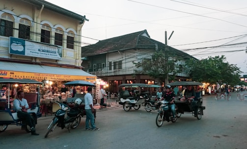A busy street scene in an Asian city with several tuk-tuks and motorcycles parked and moving. On the left, there is an art shop displaying various colorful items under a yellow sign. Many people are gathered around, with some appearing as tourists and others as locals. Buildings with traditional architecture and a mix of old and new styles surround the street.