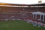 A crowded football stadium filled with passionate fans, many wearing red, celebrating and supporting their team with banners and flags. The atmosphere is lively, with a match taking place on the field, and the stadium is decorated with various colorful signs and banners.