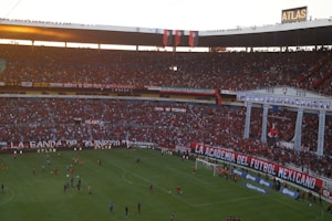 A crowded football stadium filled with passionate fans, many wearing red, celebrating and supporting their team with banners and flags. The atmosphere is lively, with a match taking place on the field, and the stadium is decorated with various colorful signs and banners.