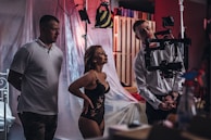 Three people are in a studio setting, with one operating a camera on a stabilizer. The woman, in a black lace outfit, appears to be the subject of the shoot, looking confident. Behind them, props and lighting equipment are set up for a photo or video shoot.