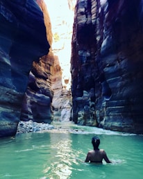 A person is wading through a narrow canyon with steep, high walls made of layered rock formations. The water has a greenish hue, and sunlight is casting dramatic shadows and highlights on the rock surfaces.