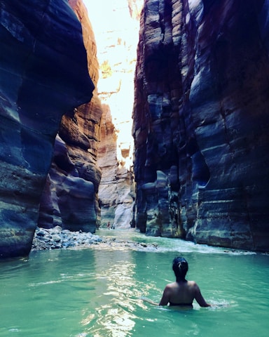 A person is wading through a narrow canyon with steep, high walls made of layered rock formations. The water has a greenish hue, and sunlight is casting dramatic shadows and highlights on the rock surfaces.
