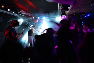 A lively concert scene with a band performing on stage under vibrant lighting. The stage is filled with a mix of smoke and colorful lights, creating an energetic atmosphere. A guitarist, a singer, and possibly a drummer are visible, along with audience members enthusiastically enjoying the music.