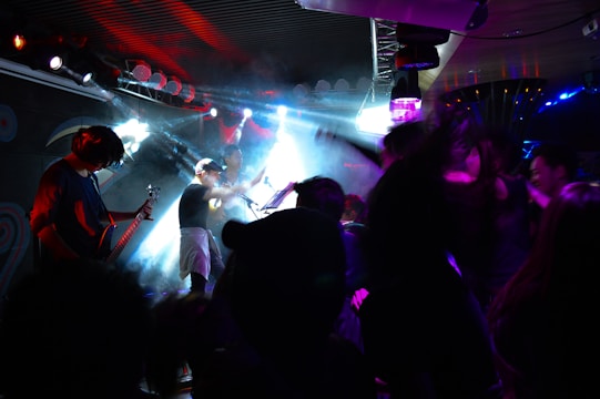 A lively concert scene with a band performing on stage under vibrant lighting. The stage is filled with a mix of smoke and colorful lights, creating an energetic atmosphere. A guitarist, a singer, and possibly a drummer are visible, along with audience members enthusiastically enjoying the music.