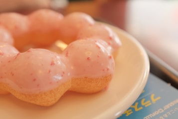 A close-up of a donut with a glossy pink frosting featuring red specks, displayed on a white plate. The background includes a blurred blue surface with yellow text.