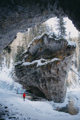 person in red jacket and black pants standing on snow covered ground near gray rocky mountain