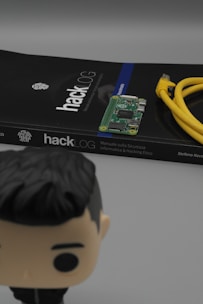 A book titled 'hackLOG' is prominently placed, accompanied by a small circuit board on top and a yellow Ethernet cable coiled beside it. In the foreground, a blurred figure of a small toy or figurine with dark hair is partially visible.