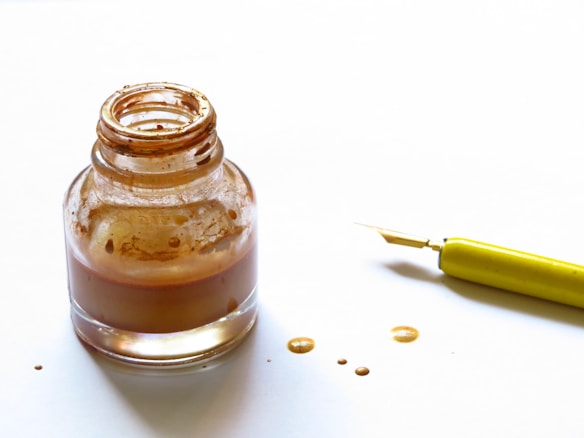 An open glass ink bottle with brown ink stains surrounding it, accompanied by a fountain pen with a yellow handle on a white surface.