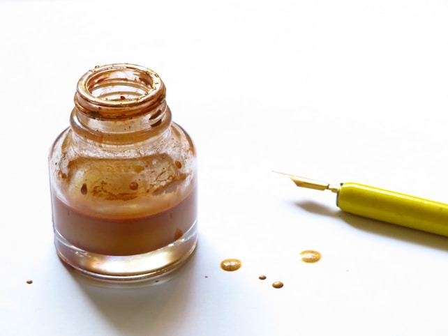 An open glass ink bottle with brown ink stains surrounding it, accompanied by a fountain pen with a yellow handle on a white surface.