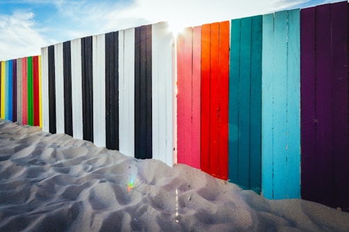 A vibrant fence composed of vertical wooden planks painted in a variety of colors, including black, white, red, blue, and purple, stands on sandy ground. The sun peeks through a gap between the planks, casting a soft light across the scene. The sand appears smooth and untouched, creating a peaceful atmosphere.