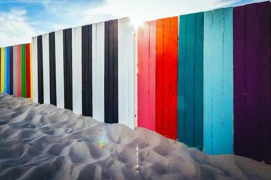 A vibrant fence composed of vertical wooden planks painted in a variety of colors, including black, white, red, blue, and purple, stands on sandy ground. The sun peeks through a gap between the planks, casting a soft light across the scene. The sand appears smooth and untouched, creating a peaceful atmosphere.