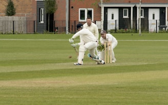 A cricket match is taking place on a grassy field. A batsman is wearing protective gear, including a helmet, gloves, and pads, and is attempting to hit the ball. Two fielders, one in wicketkeeper gear, are positioned behind the stumps, watching the ball closely. Residential buildings are seen in the background.