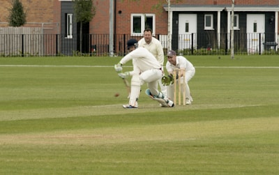 A cricket match is taking place on a grassy field. A batsman is wearing protective gear, including a helmet, gloves, and pads, and is attempting to hit the ball. Two fielders, one in wicketkeeper gear, are positioned behind the stumps, watching the ball closely. Residential buildings are seen in the background.