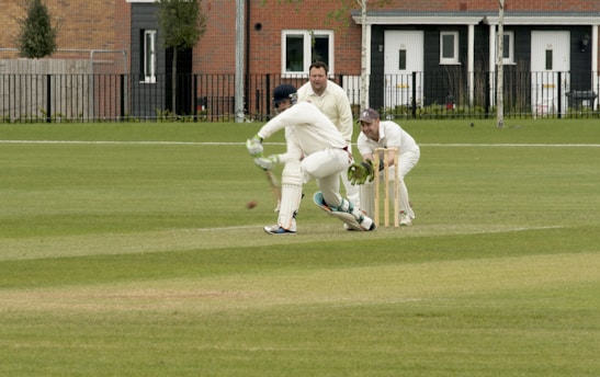 A cricket match is taking place on a grassy field. A batsman is wearing protective gear, including a helmet, gloves, and pads, and is attempting to hit the ball. Two fielders, one in wicketkeeper gear, are positioned behind the stumps, watching the ball closely. Residential buildings are seen in the background.