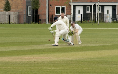 A cricket match is taking place on a grassy field. A batsman is wearing protective gear, including a helmet, gloves, and pads, and is attempting to hit the ball. Two fielders, one in wicketkeeper gear, are positioned behind the stumps, watching the ball closely. Residential buildings are seen in the background.