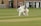 A cricket match is taking place on a grassy field. A batsman is wearing protective gear, including a helmet, gloves, and pads, and is attempting to hit the ball. Two fielders, one in wicketkeeper gear, are positioned behind the stumps, watching the ball closely. Residential buildings are seen in the background.