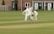 A cricket match is taking place on a grassy field. A batsman is wearing protective gear, including a helmet, gloves, and pads, and is attempting to hit the ball. Two fielders, one in wicketkeeper gear, are positioned behind the stumps, watching the ball closely. Residential buildings are seen in the background.