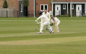 A cricket match is taking place on a grassy field. A batsman is wearing protective gear, including a helmet, gloves, and pads, and is attempting to hit the ball. Two fielders, one in wicketkeeper gear, are positioned behind the stumps, watching the ball closely. Residential buildings are seen in the background.