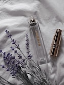 A clear perfume bottle with gold accents is placed on a soft white fabric, accompanied by a gold-capped lipstick and sprigs of lavender. The setting suggests a subtle, elegant atmosphere.